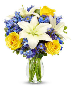 Yellow roses with blue delphinium and white lilies in a clear glass vase